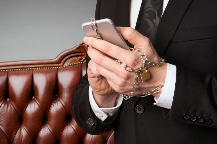 Homem de terno com as mãos acorrentadas ao celular, segurando-o enquanto um cadeado dourado prende a corrente. A imagem sugere uma dependência, talvez em relação à tecnologia ou uma alusão à ludopatia, com um sofá de couro em segundo plano.