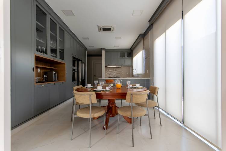 Uma cozinha moderna e elegante com armários cinza. Há uma mesa de jantar redonda de madeira com quatro cadeiras estofadas claras no centro. Ao fundo, uma janela grande com persianas.