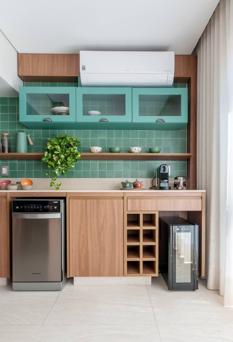 Uma cozinha moderna e colorida. Armários de madeira clara e três armários superiores em azul-petróleo. A parede possui azulejos verdes e prateleiras de madeira. Há uma lava-louças de aço inoxidável e uma adega de vinho compacta.