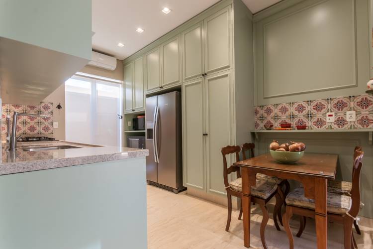 Uma cozinha com armários verde-claro e bancada cinza. Há uma geladeira de aço inoxidável e uma área de jantar com mesa de madeira escura e cadeiras. A parede sobre a mesa possui azulejos decorativos e um prato de frutas.