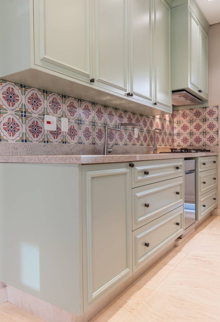 Uma cozinha charmosa com armários verde-claro. O destaque é o backsplash com azulejos coloridos e padrões geométricos. Há uma bancada cinza e uma pia de aço inoxidável. Eletrodomésticos embutidos complementam o design.