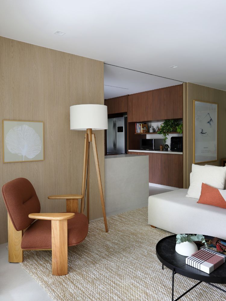 Parte do living com poltrona marrom, luminária de chão, sofá orgânico branco, tapete bege e cozinha ao fundo