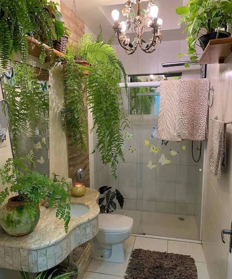 Um banheiro exuberante com diversas plantas penduradas e em vasos, incluindo samambaias volumosas. Um lustre ornamentado ilumina o espaço, que possui um balcão de mármore e detalhes de borboletas na parede do chuveiro, criando um oásis verde.