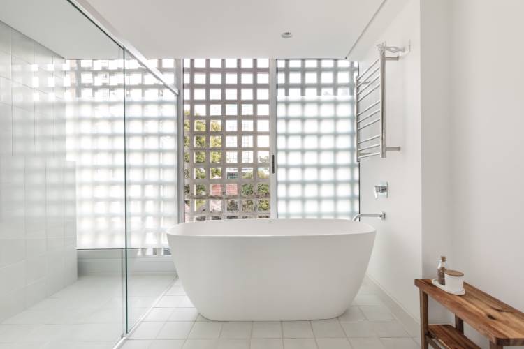 Apartamento-Reforma-Compelta-12 Um banheiro contemporâneo com uma banheira branca oval central. A parede dos fundos é composta por tijolos de vidro que permitem a entrada de luz e privacidade. Um box de vidro está à esquerda, e uma estante de madeira rústica e um toalheiro aquecido completam o ambiente.