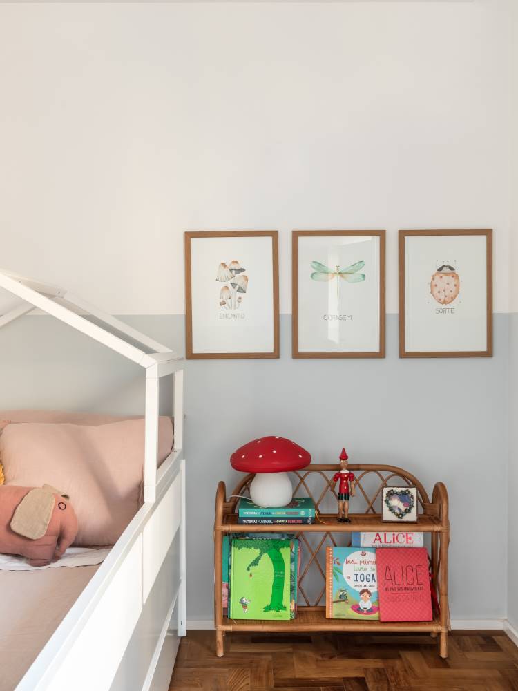 Um quarto infantil aconchegante com uma cama em formato de casinha. As paredes são brancas e cinza claro, decoradas com quadros de ilustrações. Uma estante de vime com livros e uma luminária de cogumelo vermelho adicionam um toque lúdico ao espaço.