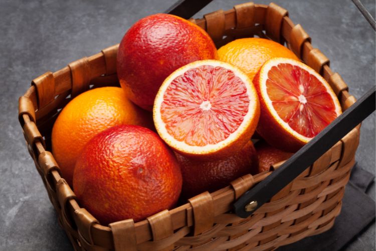 Uma cesta de vime marrom cheia de laranjas de corte sanguíneo inteiras e cortadas ao meio, exibindo sua polpa vibrante e avermelhada, está sobre uma superfície escura.