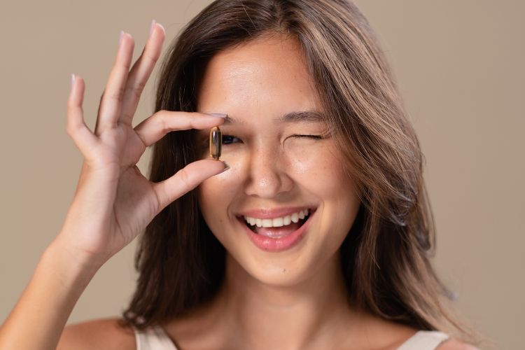 Uma mulher jovem, com cabelo castanho escuro e um sorriso radiante, pisca um olho enquanto segura uma cápsula marrom-dourada entre os dedos, posicionando-a na frente do outro olho, contra um fundo neutro.