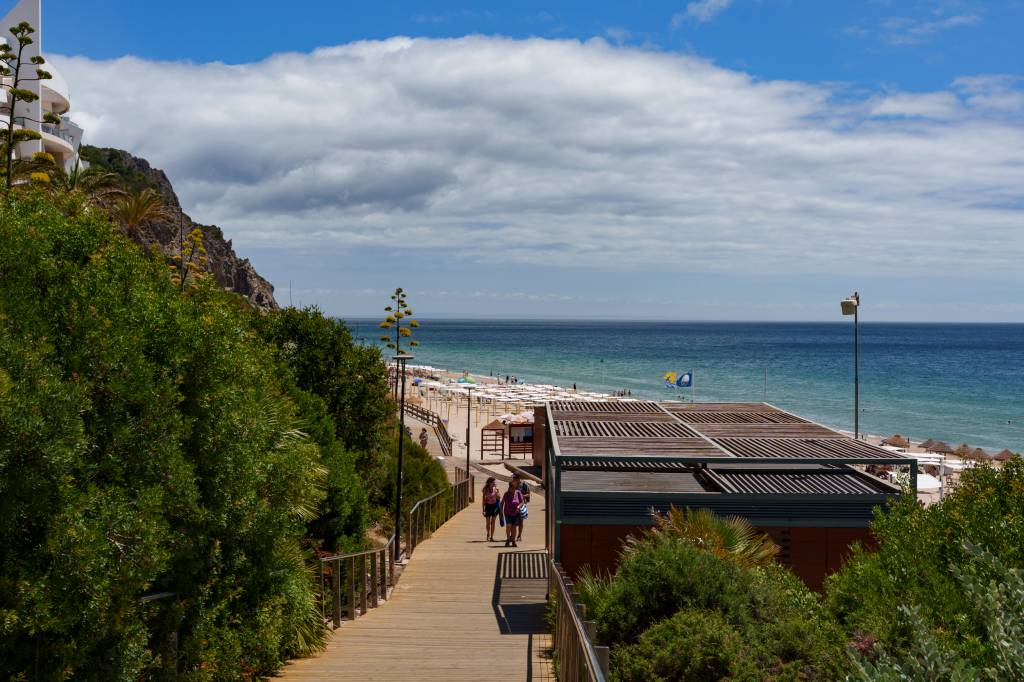 Uma passarela de madeira leva turistas ate a areia da praia, com uma densa vegetação verde circundando