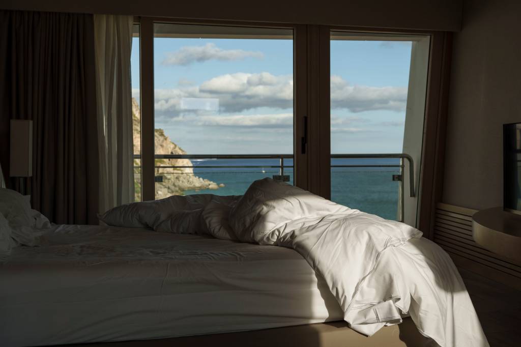 Cama desarrumada num quarto de hotel, com uma varanda de vidro que revela o mar la fora