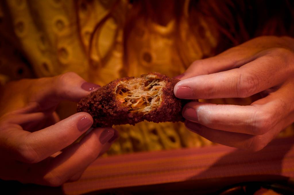 Imagem fechada nas mãos de uma mulher, que abre um croquete para comê-lo