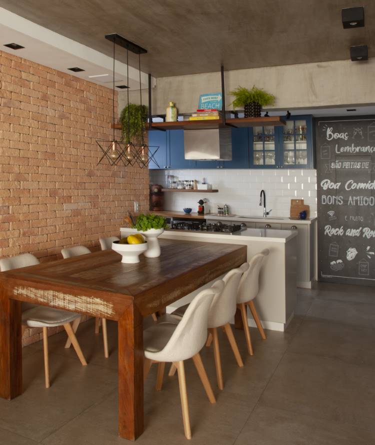 Cozinha-Rustica-Moderna-9 Cozinha e sala de jantar integradas com parede de tijolinhos e teto de cimento queimado. A mesa de madeira rústica com cadeiras brancas modernas contrasta com a cozinha de armários azuis, criando um ambiente contemporâneo.
