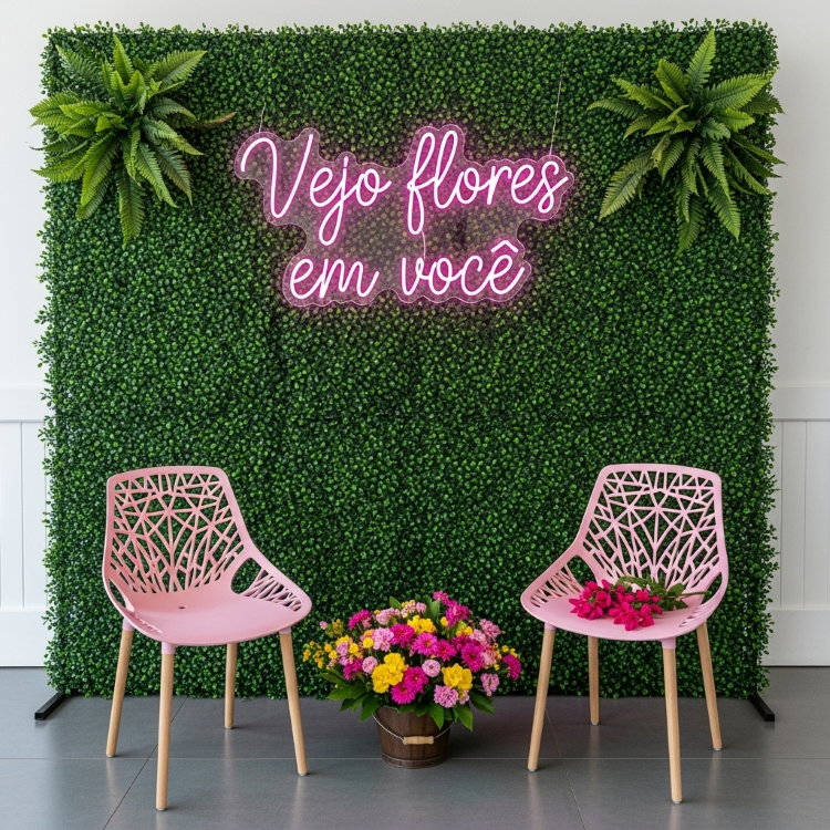 Uma parede coberta por folhagem artificial e decorada com flores falsas. Na frente, duas cadeiras rosas com pernas de madeira. Entre elas, um balde de flores. Acima, um letreiro de neon rosa diz "Vejo flores em você".