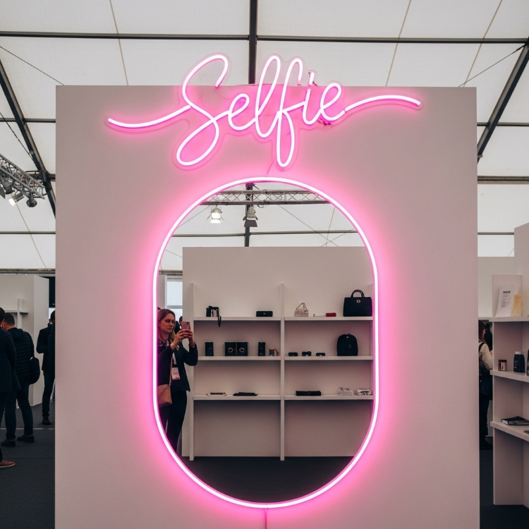 Uma pessoa tira uma foto de si mesma na frente de um espelho com formato oval e contorno de neon rosa. Acima, um letreiro de neon rosa diz "Selfie". Há prateleiras brancas com bolsas e carteiras atrás do espelho.