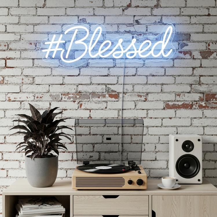 Um close de um toca-discos de madeira bege tocando um vinil. À esquerda há um vaso de plantas escuro, à direita uma xícara de café e uma caixa de som branca. Acima, em uma parede de tijolos brancos, uma luz de neon azul diz "#Blessed".