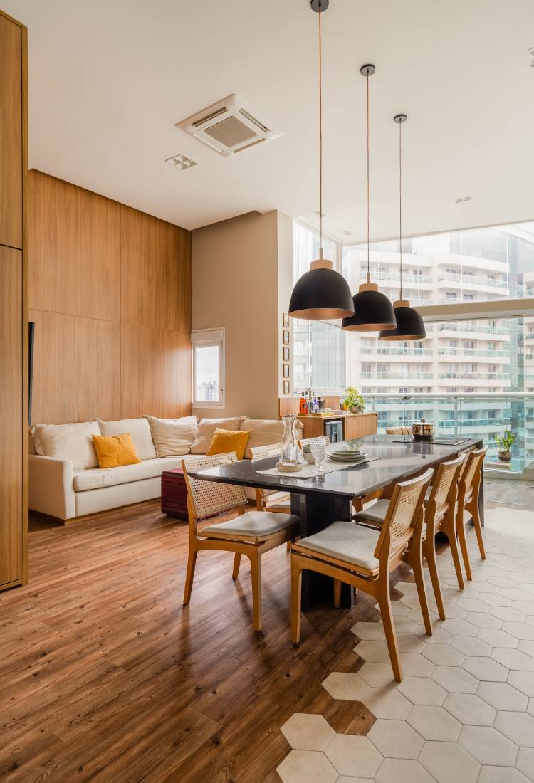 Sala de estar e jantar integrada. A área de jantar, com piso de ladrilhos hexagonais, tem uma mesa preta para seis pessoas, com cadeiras de madeira e palha. Acima, três luminárias pretas pendentes. A sala de estar, com piso de madeira, tem um sofá branco e painel de madeira na parede.