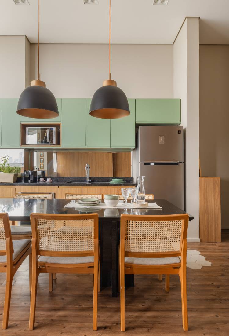 Cozinha moderna com armários superiores em verde-claro e inferiores em madeira. Uma ilha preta de jantar com cadeiras de madeira e palha no primeiro plano. Duas luminárias pendentes pretas com interior dourado iluminam a ilha. O piso é de madeira e o teto é branco com iluminação embutida.