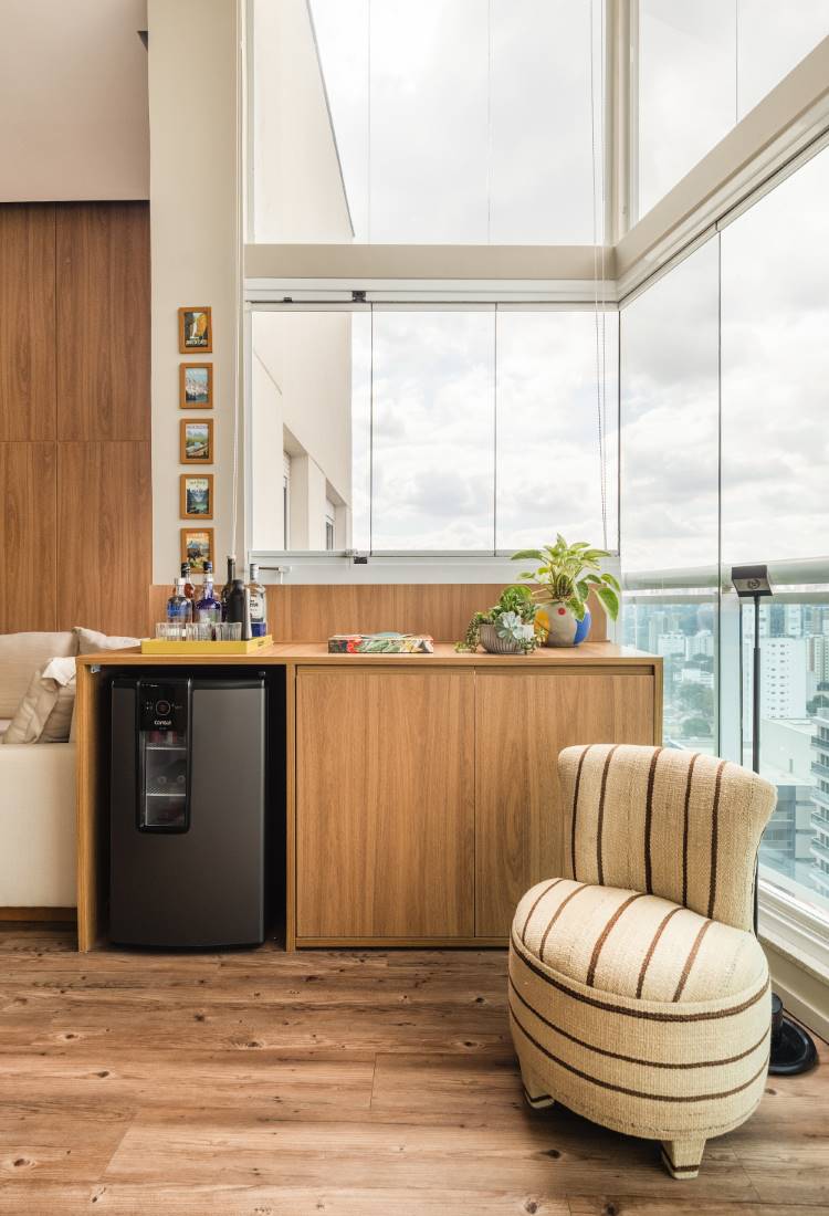 Varanda gourmet moderna com vista para a cidade. Um balcão de madeira com armários e uma cervejeira preta embutida. Em cima do balcão, bebidas, objetos decorativos e uma planta. À direita, uma poltrona listrada e, ao fundo, janelas de vidro.