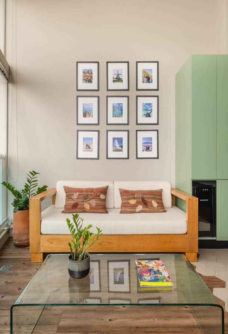 Sala de estar com sofá de madeira e almofadas estampadas. Acima do sofá, uma galeria de parede com nove quadros. Na frente, uma mesa de centro de vidro com um vaso de planta e um livro. Ao lado, um armário verde claro e um vaso de planta grande.