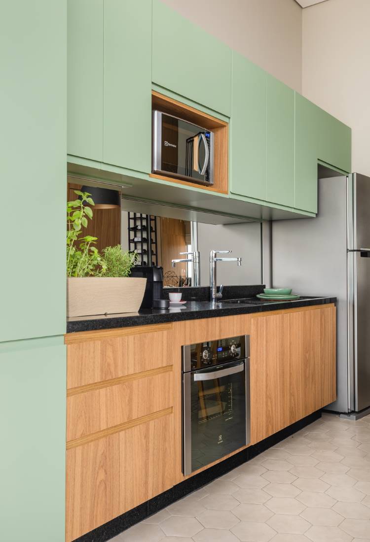 Cozinha moderna com armários superiores em verde-claro e inferiores em madeira. Micro-ondas e forno elétrico embutidos. Bancada preta com pia, torneira e uma planta. O backsplash espelhado reflete a varanda. O piso tem ladrilhos hexagonais brancos, e a geladeira de inox aparece ao lado.