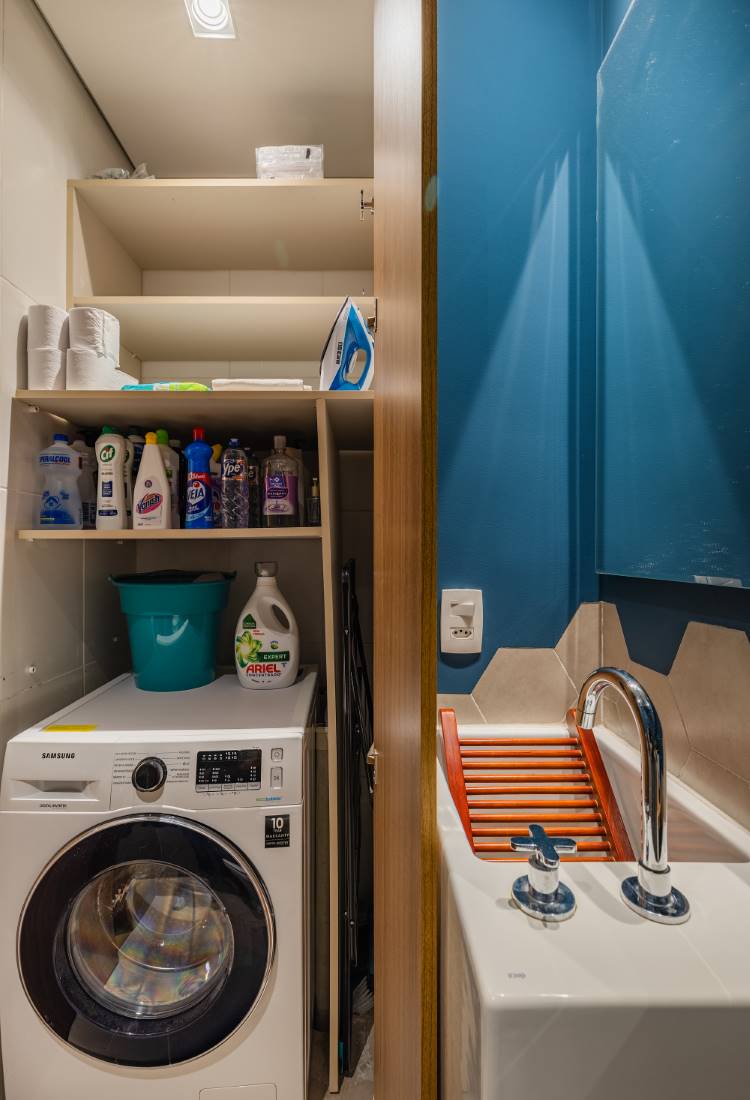 Área de serviço compacta com uma máquina de lavar roupa branca. Acima, prateleiras abertas com produtos de limpeza e um ferro de passar. Ao lado, um tanque branco com torneira metálica e um painel de madeira. A parede é azul e o piso é branco.
