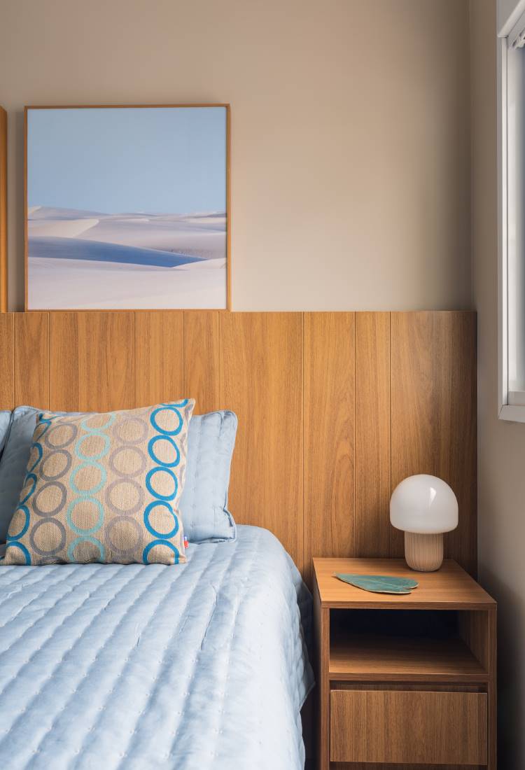 Quarto moderno de um estúdio de 77 m². A imagem mostra uma cama com cabeceira de madeira e roupa de cama azul. A cama tem uma almofada decorativa de estampa geométrica. Na parede, um quadro com a paisagem dos lençóis. Ao lado, um criado-mudo com abajur branco.