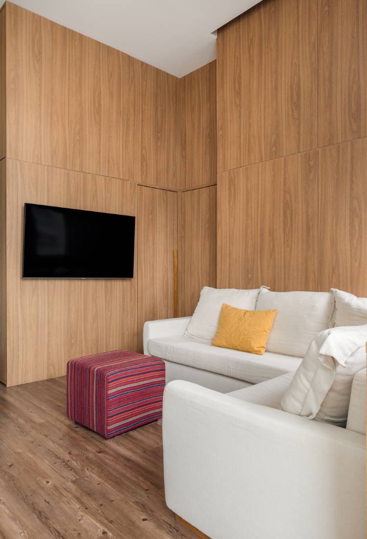 Sala de estar de um estúdio de 77 m². A imagem mostra um painel de madeira com TV preta embutida, criando um ambiente minimalista e moderno. A sala tem dois sofás brancos com almofadas, uma amarela e uma listrada, e um puff listrado roxo. O piso é de madeira.