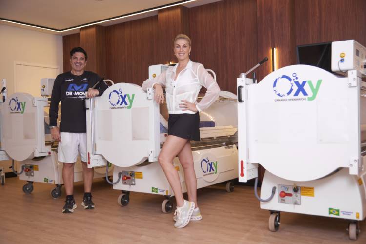 Ana Hickmann e Edu Guedes posam em uma clínica ao lado de várias câmaras hiperbáricas brancas com o logo azul "Oxy". O homem usa uma camiseta preta "DR MOVE" e a mulher, uma blusa branca e shorts pretos.