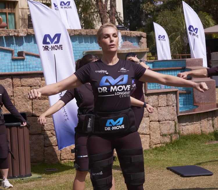 Ana Hickmann com uma camiseta preta "DR MOVE", pratica exercícios com os braços estendidos. Ela usa um cinto de eletroestimulação e faixas nas pernas, com bandeiras brancas da "DR MOVE" ao fundo.