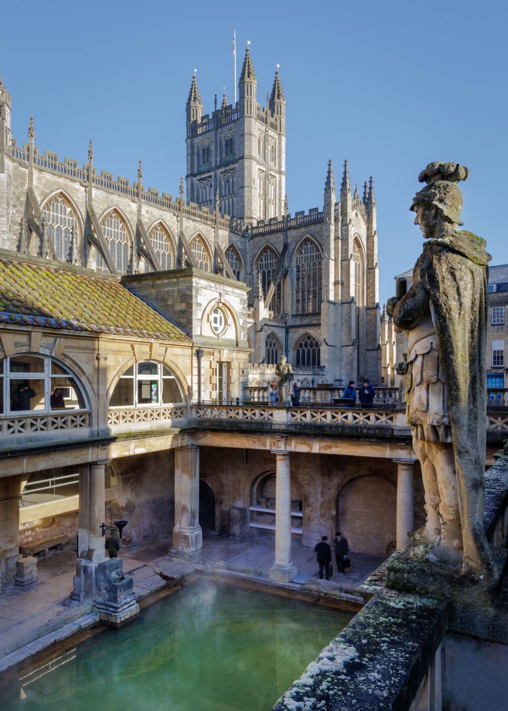 A Large Sculpture Stands On Top Of Roman Baths Ruins With An Abbey Beyond termas romanas inglaterra