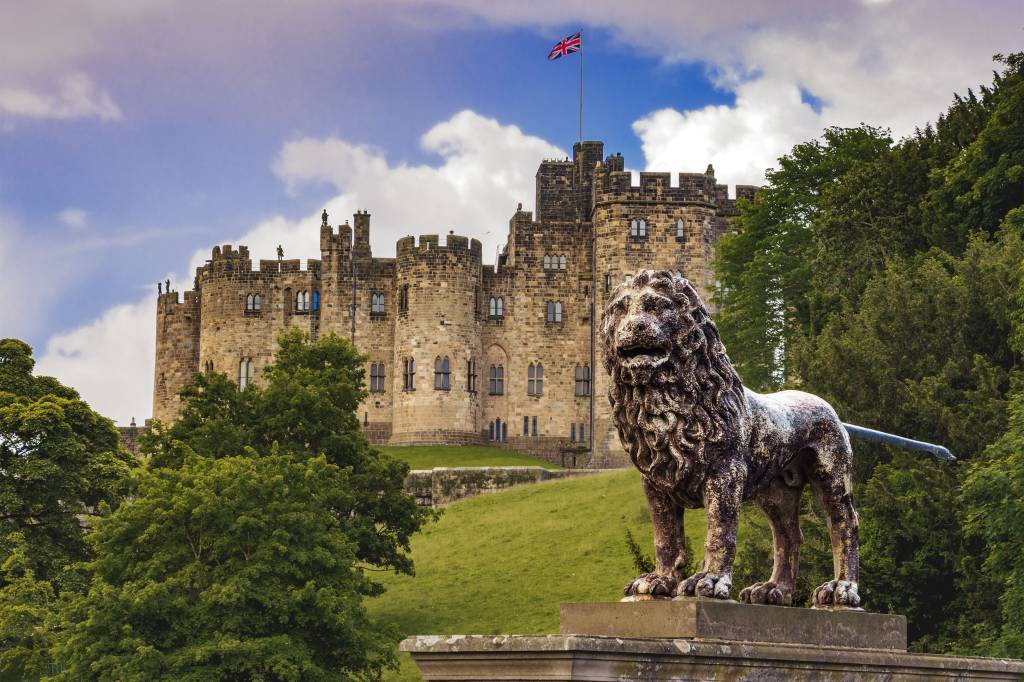 An Historic Castle With A Uk Flag At Full Mast And With A Lion Statue Out The Front. Alnwick Castle