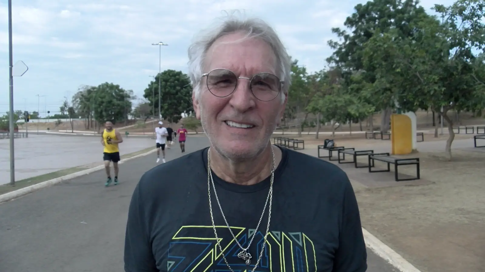 00030000-0Aa4-0A58-1F48-08De12664F39 Homem de cabelos grisalhos e óculos sorri durante corrida em parque arborizado, usando camiseta escura e colares prateados.