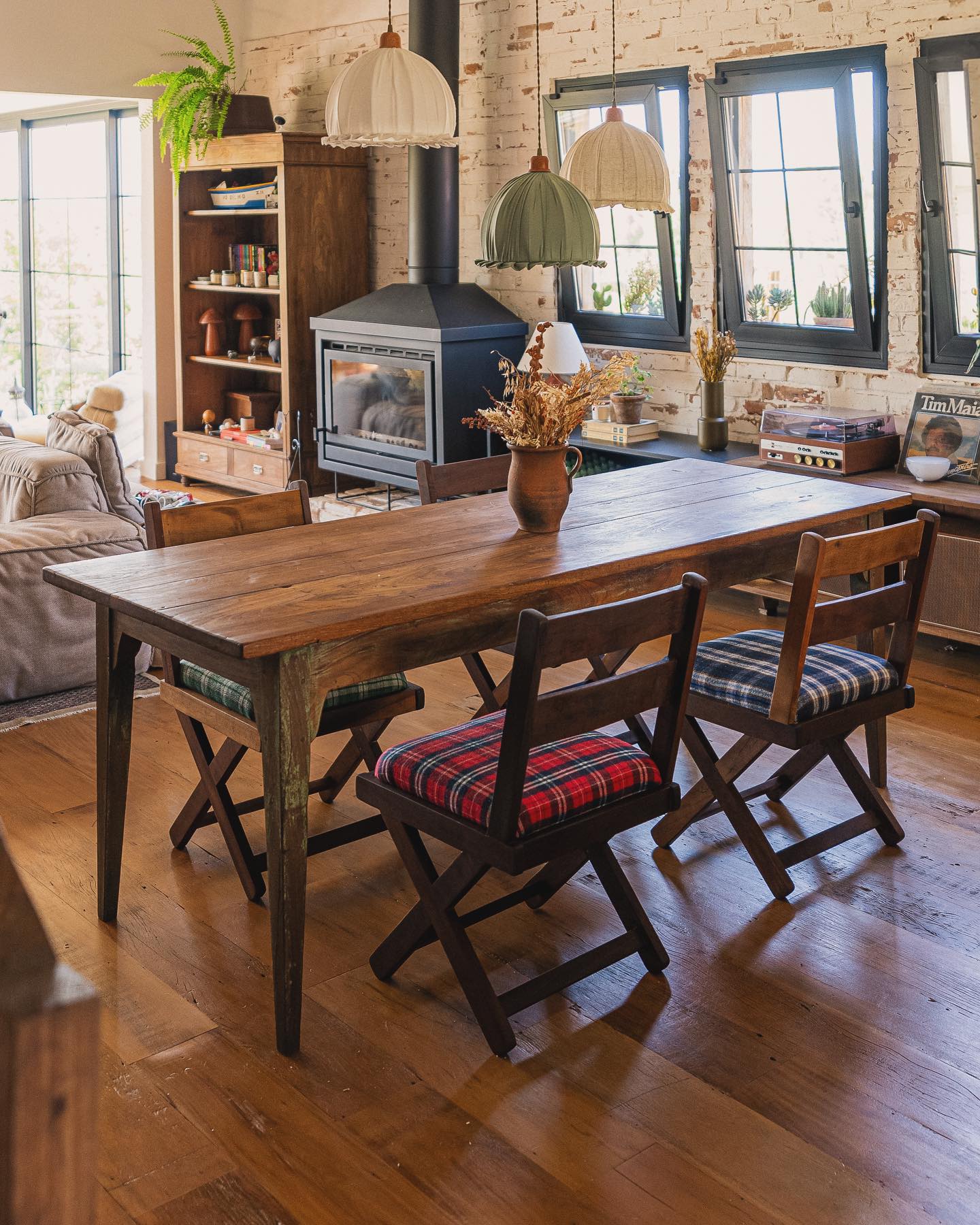 Decoração de sala de jantar farmhouse com mesa rústica, cadeiras com estofado xadrez e luminárias de tecido.