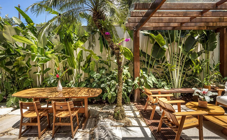 Quintal com mesa e cadeiras de madeira cercadas por plantas tropicais, mostrando como o verde traz frescor e aconchego ao ambiente externo.