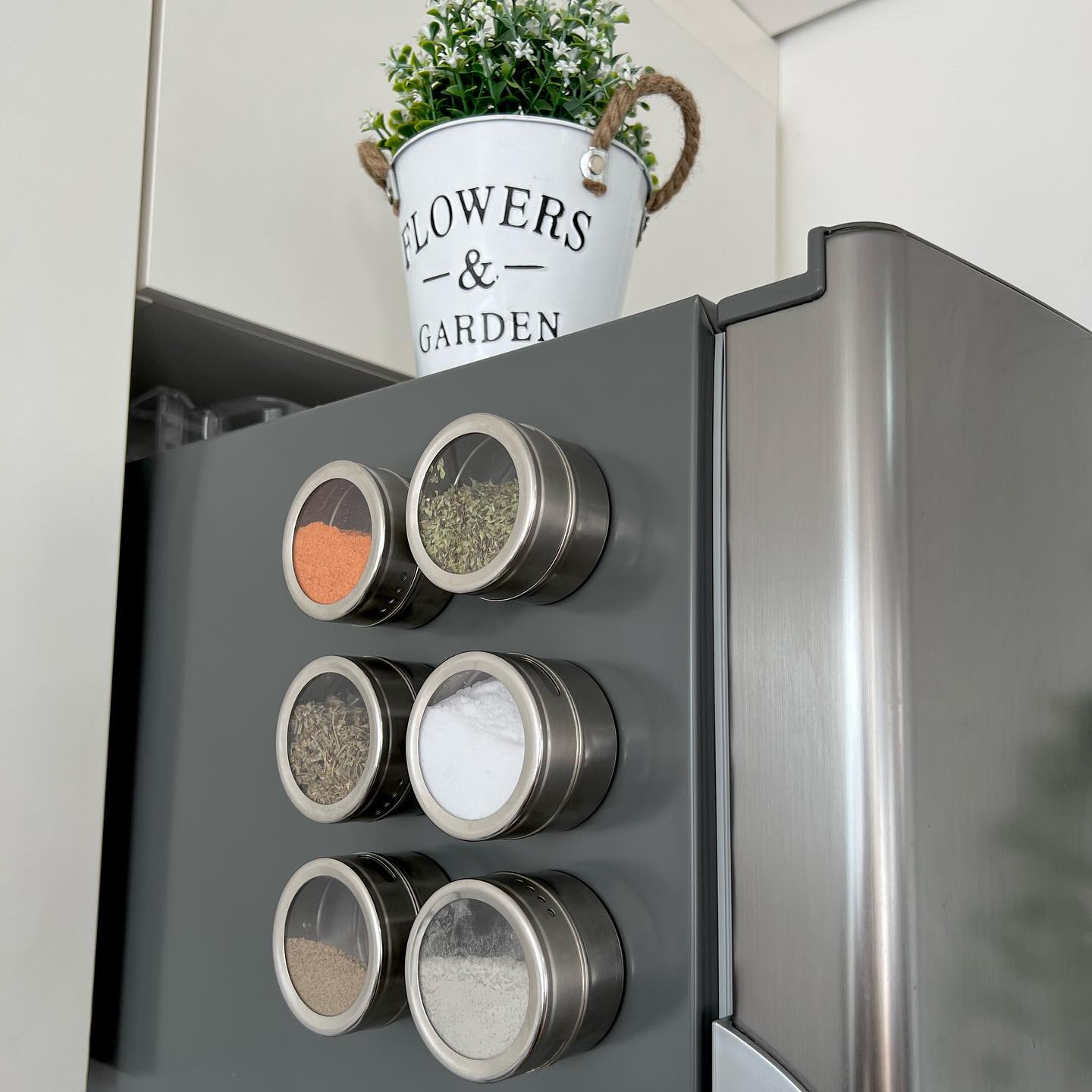 Recipientes magnéticos de temperos fixados na lateral da geladeira em cozinha moderna, mostrando solução prática de organização e aproveitamento de espaço.