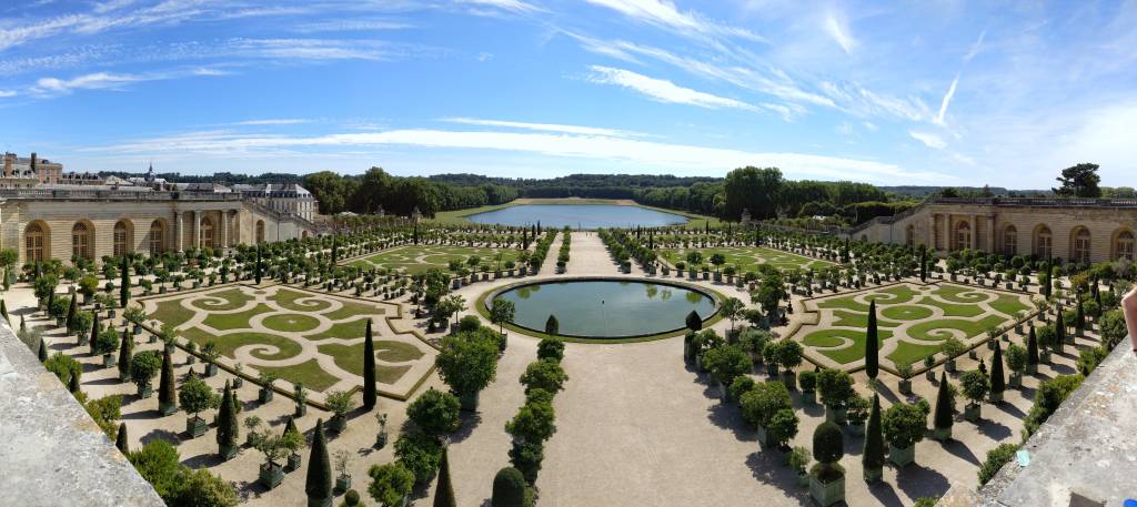 Vista panorâmica de jardins com grama desenhada, árvores alinhadas e plantadas de forma coordenada, dois lados e um palácio à esquerda