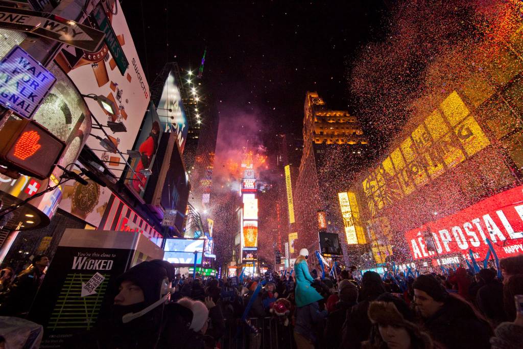 Multidão na Times Square, rua com prédios altos, coloridos com anúncios. Confeti cai do céu.