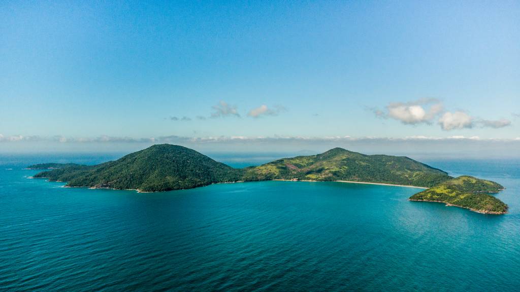 Vista aérea de ilha, coberta por vegetação e cercada por mar de cor azulada.