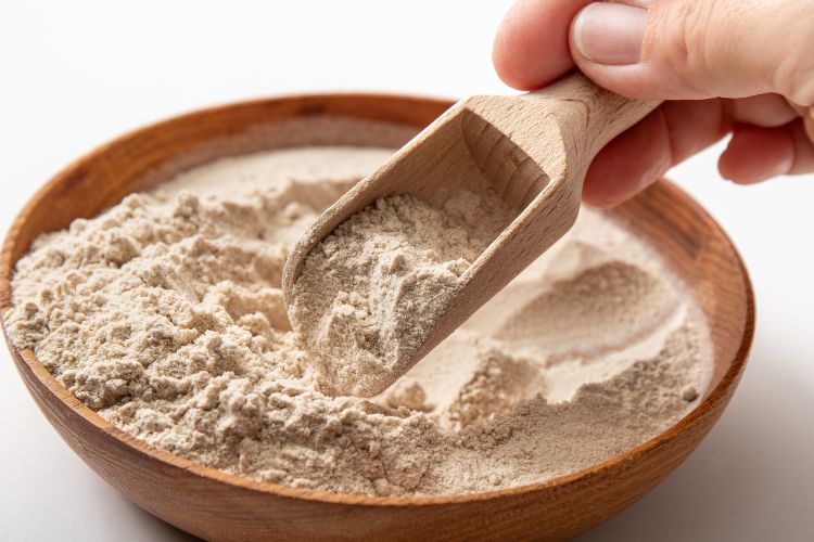 Psyllium-Para-Emagrecer-3 A imagem mostra uma tigela de madeira cheia de psyllium em pó. Uma mão segura uma colher de madeira, retirando um pouco do pó da tigela. O fundo é neutro e claro, destacando a textura fina e a cor bege do psyllium. A cena é simples e focada nos detalhes do ingrediente natural.