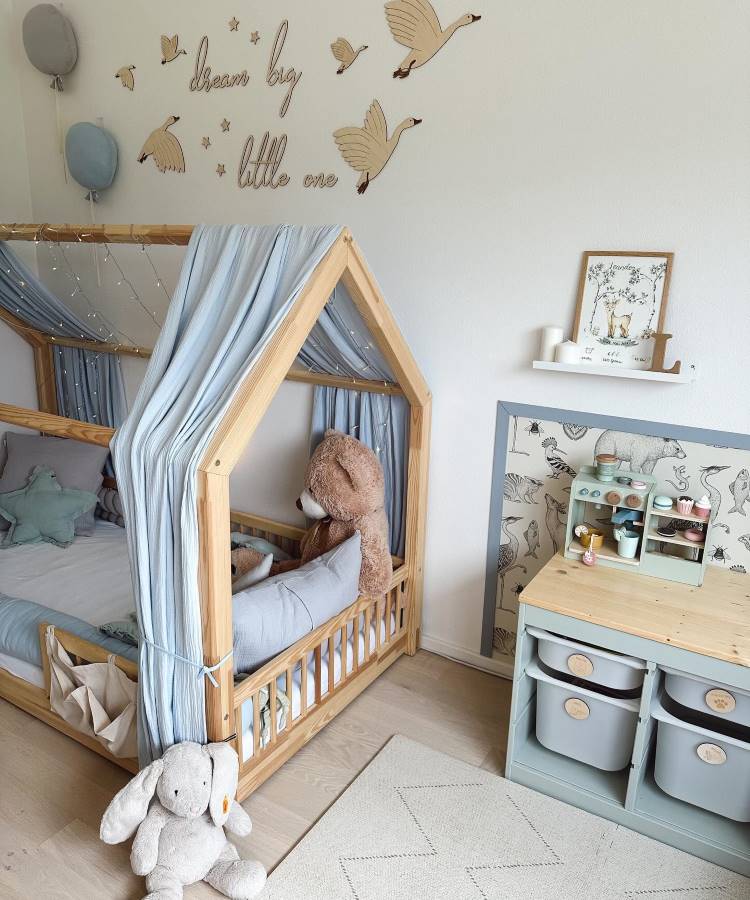 Dicas-Decoracao-Para-Quem-Tem-Tdah-8 O quarto infantil apresenta uma cama em formato de casinha de madeira com cortinas azul-claro e luzes decorativas. Um grande urso de pelúcia está sobre a cama. Na parede, adesivos de gansos voando e a frase “dream big little one”. Uma estante organiza brinquedos e utensílios.