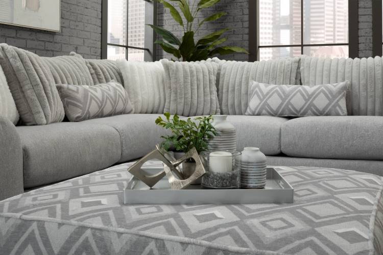 Dicas-Decoracao-Para-Quem-Tem-Tdah-2 A imagem mostra uma sala moderna com sofá cinza em "L", decorado com almofadas texturizadas e geométricas em tons neutros. Sobre o puff central, há uma bandeja prateada com velas, vasos decorativos e uma planta. Grandes janelas iluminam o ambiente.