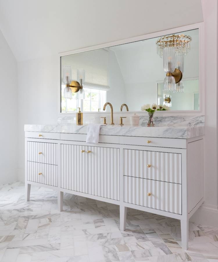 Dicas-Decoracao-Para-Quem-Tem-Tdah-4 A imagem apresenta um banheiro sofisticado com gabinete branco de ripas e puxadores dourados. A pia de mármore possui torneiras douradas elegantes. Um grande espelho reflete a luminária de cristal pendente. O piso é de mármore em tons claros.