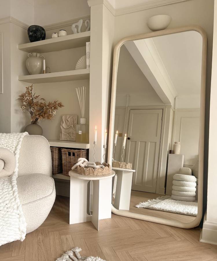 Dicas-Decoracao-Para-Quem-Tem-Tdah-13 A imagem mostra um ambiente acolhedor em tons neutros, com destaque para um espelho grande de moldura bege encostado na parede. Há uma poltrona confortável com manta de tricô, mesa lateral com cestos e velas, e prateleiras decoradas com cerâmicas e livros. O piso de madeira clara complementa o espaço.