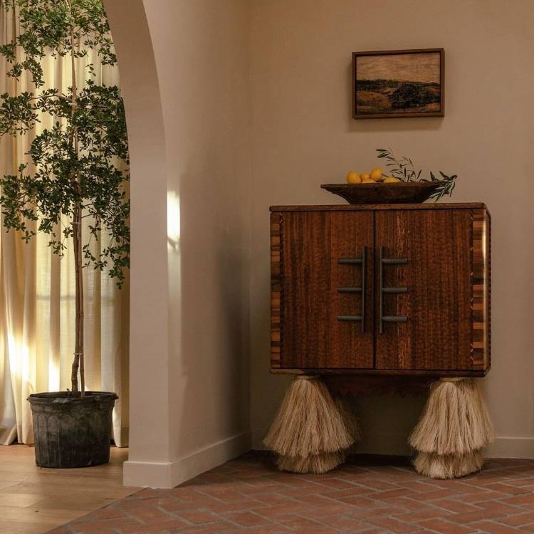 Dicas-Decoracao-Para-Quem-Tem-Tdah-16 A imagem apresenta um ambiente minimalista e rústico. Um armário de madeira com detalhes únicos em palha nas bases destaca-se, decorado com uma tigela de madeira contendo limões e ramos verdes. Ao lado, uma planta em vaso metálico complementa a estética natural.