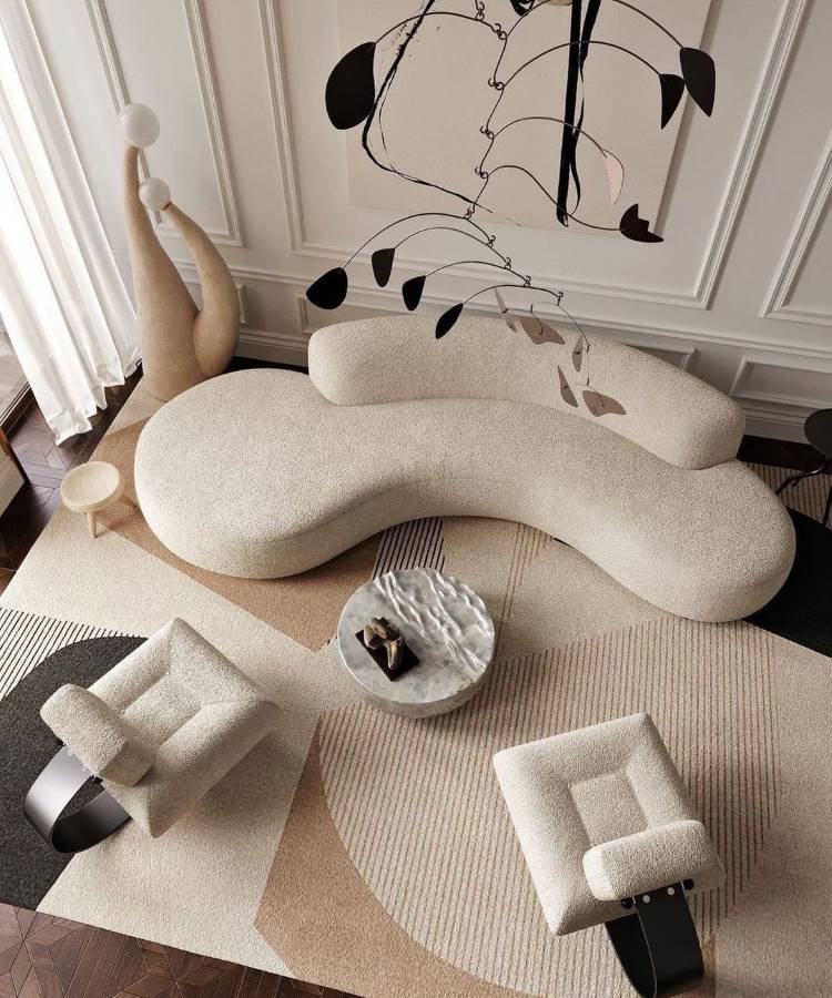 Dicas-Decoracao-Para-Quem-Tem-Tdah-5 A imagem apresenta uma sala moderna com sofá curvo bege, poltronas geométricas e uma mesa central de mármore. O tapete com padrões suaves complementa o ambiente. Dicas de decoração para quem tem TDAH incluem espaços minimalistas e móveis funcionais.