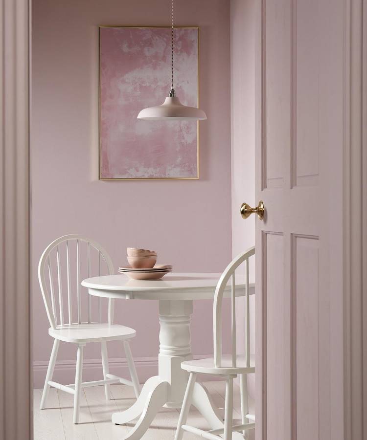 Dicas-Decoracao-Para-Quem-Tem-Tdah-6 A imagem mostra um ambiente delicado com paredes rosa claro, uma mesa redonda branca e cadeiras combinando. Um quadro abstrato rosa e luminária pendente completam o espaço. Dicas de decoração para quem tem TDAH incluem cores suaves e organização visual.