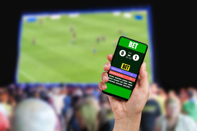 Ludopatia-Transtorno-5 Uma mão segura um smartphone exibindo um aplicativo de apostas com resultados de jogo, possivelmente simulando o Bet365. Ao fundo, uma partida esportiva é projetada para uma multidão, ilustrando o engajamento com apostas online e a potencial ligação com a ludopatia.