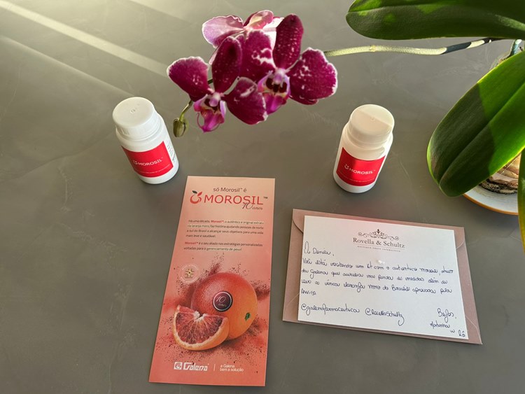 Morosil-5 Uma vista superior de dois frascos brancos de "MOROSIL" com rótulos vermelhos, um folheto informativo sobre Morosil e um cartão de agradecimento manuscrito da Rovella & Schultz. Uma orquídea roxa e folhas verdes decoram a imagem.