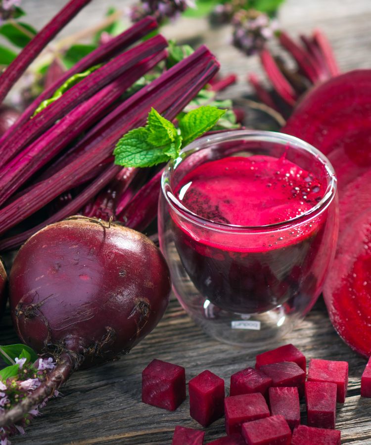 Tudo-Sobre-Beterraba-2 Um copo de suco de beterraba vibrante com uma folha de hortelã. Ao redor, há uma beterraba inteira, outra cortada ao meio, cubos de beterraba e um feixe de caules, todos em uma mesa de madeira.