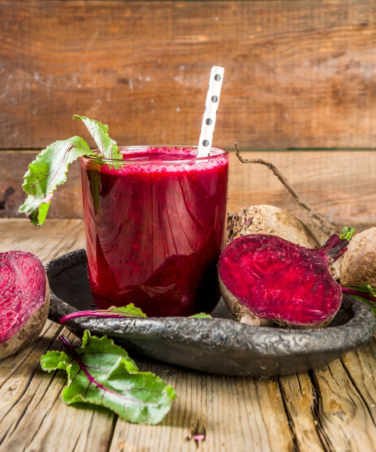 Receitas-Beterraba-1 Um close-up de um copo de suco de beterraba vermelho-escuro com um canudo. Folhas e raízes de beterraba frescas e inteiras estão em uma bandeja escura ao lado do copo, tudo sobre uma mesa de madeira rústica.
