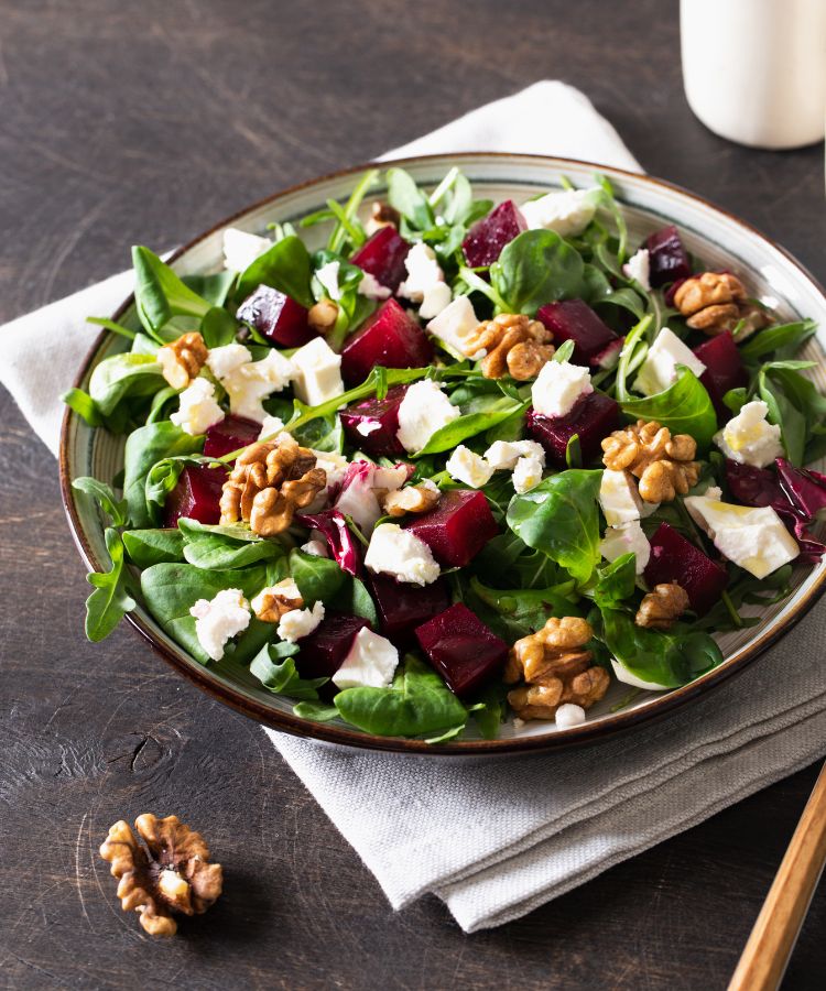 Receitas-Beterraba-2 Uma salada vibrante em um prato com folhas verdes, cubos de beterraba, nozes, e queijo feta. A salada repousa sobre um guardanapo de linho em uma mesa de madeira escura.
